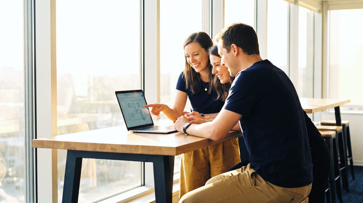 Three colleagues at a tall table by a bright window, reviewing a plan on a laptop together