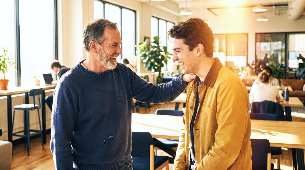 A mentor with a hand on a young man’s shoulder, both laughing in a sunlit co-working space