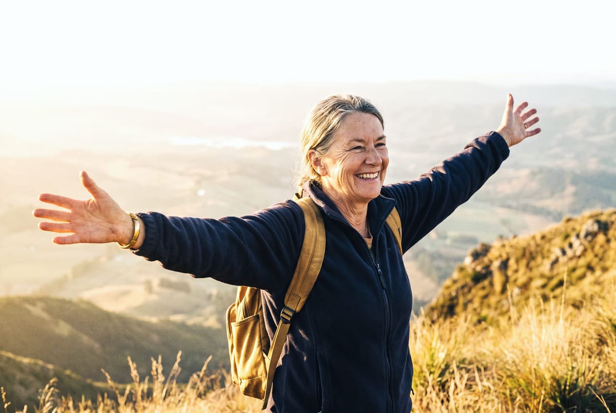 A hiker in her sixties at a mountain overlook in morning sun, arms open wide, joyful